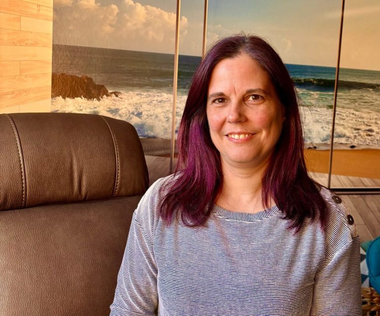 Hypnosecoachin Sonja Patermann Frau mit violetten Haaren und Brille, lächelt vor einem braunen Sessel und einem verschwommenen Hintergrund.